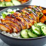 A close-up of a rice bowl topped with grilled, sliced chicken breast that has a caramelized glaze, accompanied by roasted sweet potato cubes and fresh cucumber slices. The dish is garnished with chopped parsley and served in a dark ceramic bowl on a marble surface.