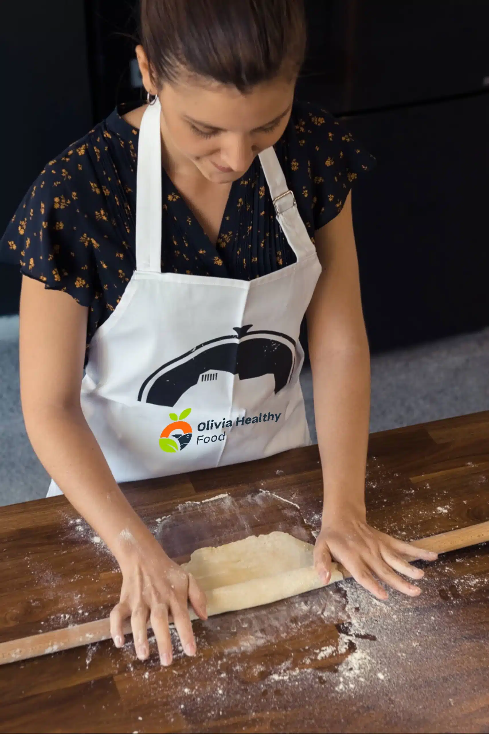 Olivia rolls out dough on a floured wooden countertop, wearing a white 'Olivia Healthy Food' apron over a black floral blouse, focused on her task