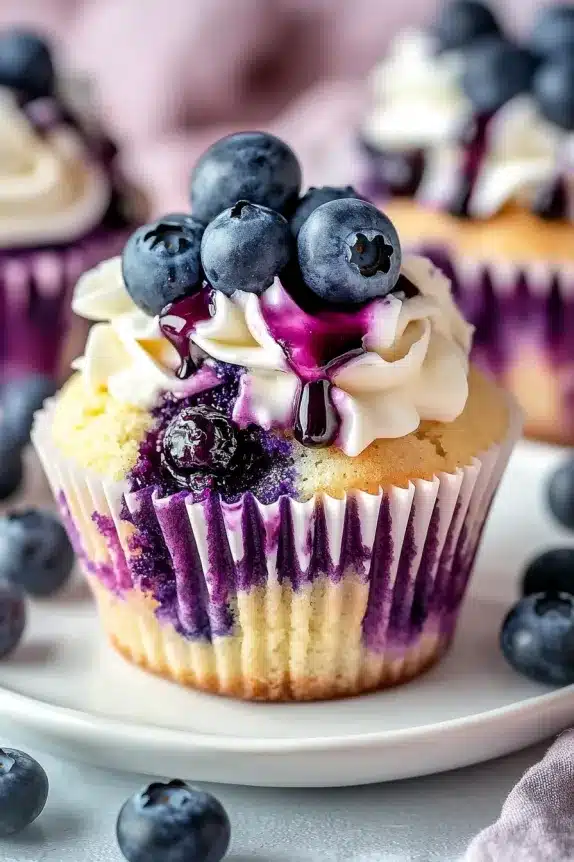 A freshly baked blueberry cupcake with vanilla frosting, topped with fresh blueberries and drizzled with blueberry sauce.