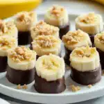 A rustic wooden board with frozen banana bites, coated in dairy-free chocolate and topped with crunchy granola and coconut flakes.