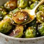 Golden-brown Brussels sprouts, glazed and topped with sesame seeds, served in a rustic bowl with a golden spoon.