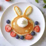 A cute bunny-shaped pancake with two ears, topped with a dollop of whipped cream, and surrounded by fresh blueberries and sliced strawberries, served on a white plate on a marble surface.