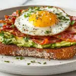 A thick slice of whole-grain toast layered with smashed avocado, crispy bacon, and a soft-boiled egg, sprinkled with sea salt and black pepper.