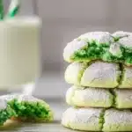 A stack of soft green crinkle cookies dusted with powdered sugar, with one cookie broken open revealing a vibrant green center, accompanied by a glass of milk with green straws.