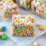 A close-up of a colorful Lucky Charms marshmallow cereal bar, featuring crispy oat cereal pieces and heart-shaped marshmallows, sitting on parchment paper with scattered cereal bits around. A fun and festive no-bake treat!