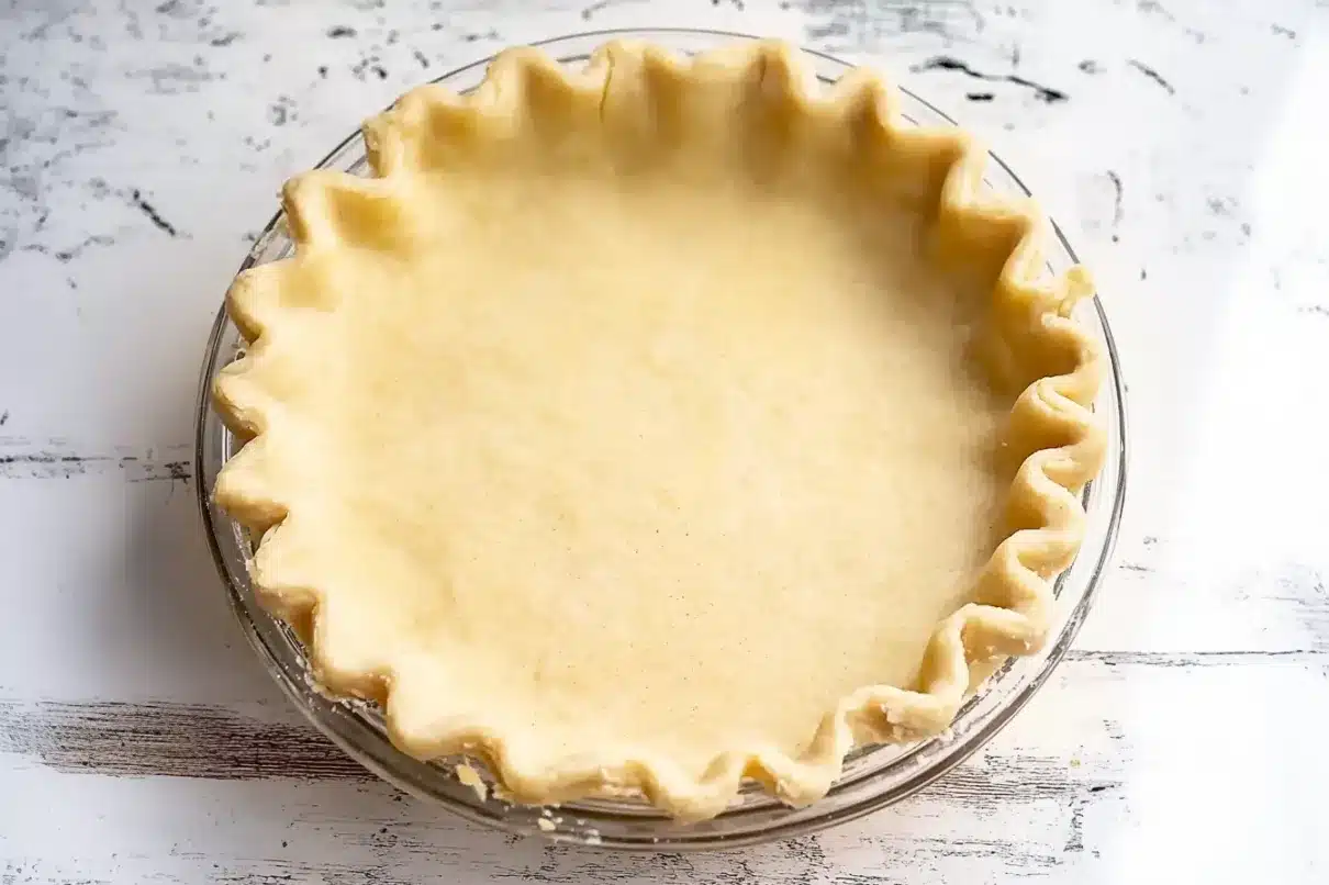 A rich, fudgy slice of chocolate chess pie on a white ceramic plate, lightly dusted with cocoa powder. The glossy chocolate filling contrasts with the crisp, golden pie crust. A fork rests beside the slice, and the whole pie is visible in the softly blurred background.