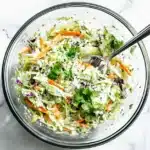 Overhead view of prepared cilantro lime slaw in a glass bowl on marble surface, featuring bright green cilantro flecks mixed with shredded cabbage and carrots.