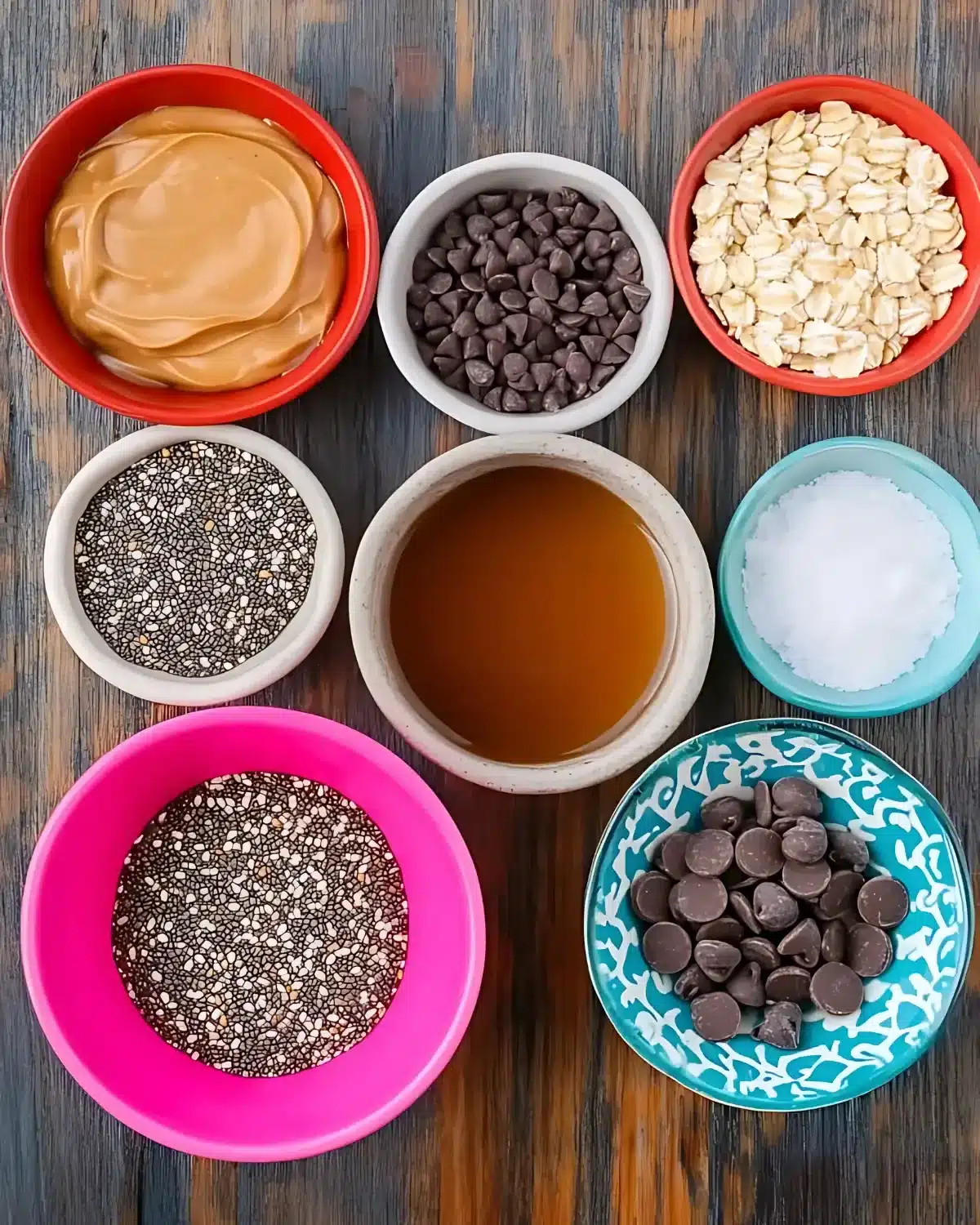 Top-down view of bowls with peanut butter, oats, chocolate chips, maple syrup, chia seeds, and salt on rustic wood
