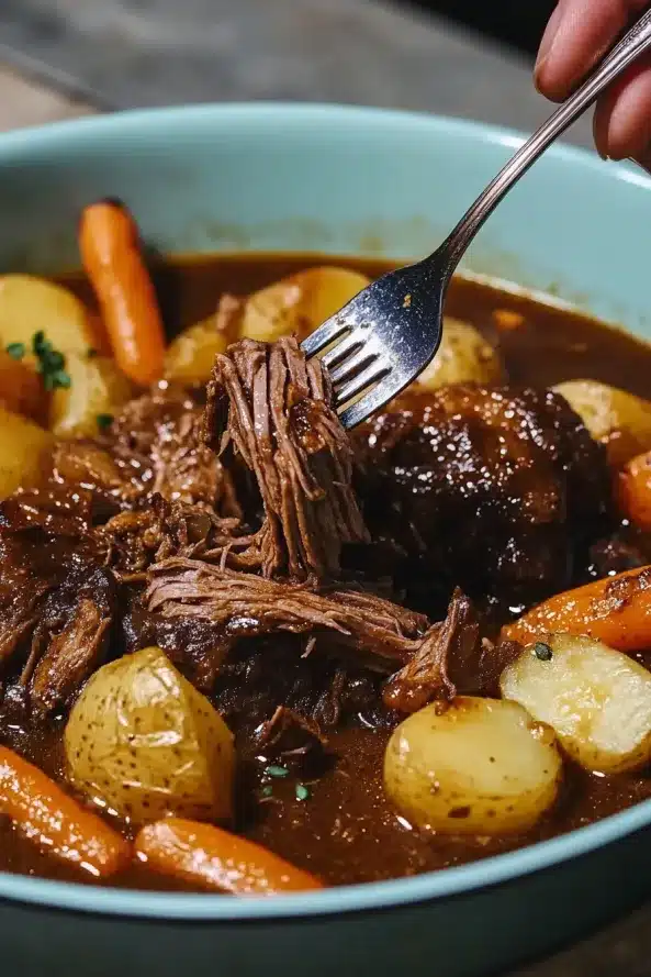 Fork lifting shredded beef from a hearty pot roast with carrots and golden potatoes in a thick brown sauce.