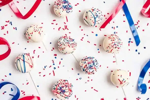 Overhead view of patriotic cake pops scattered on white surface with red and blue ribbons and sprinkles, showing various decorating styles for Independence Day celebration.
