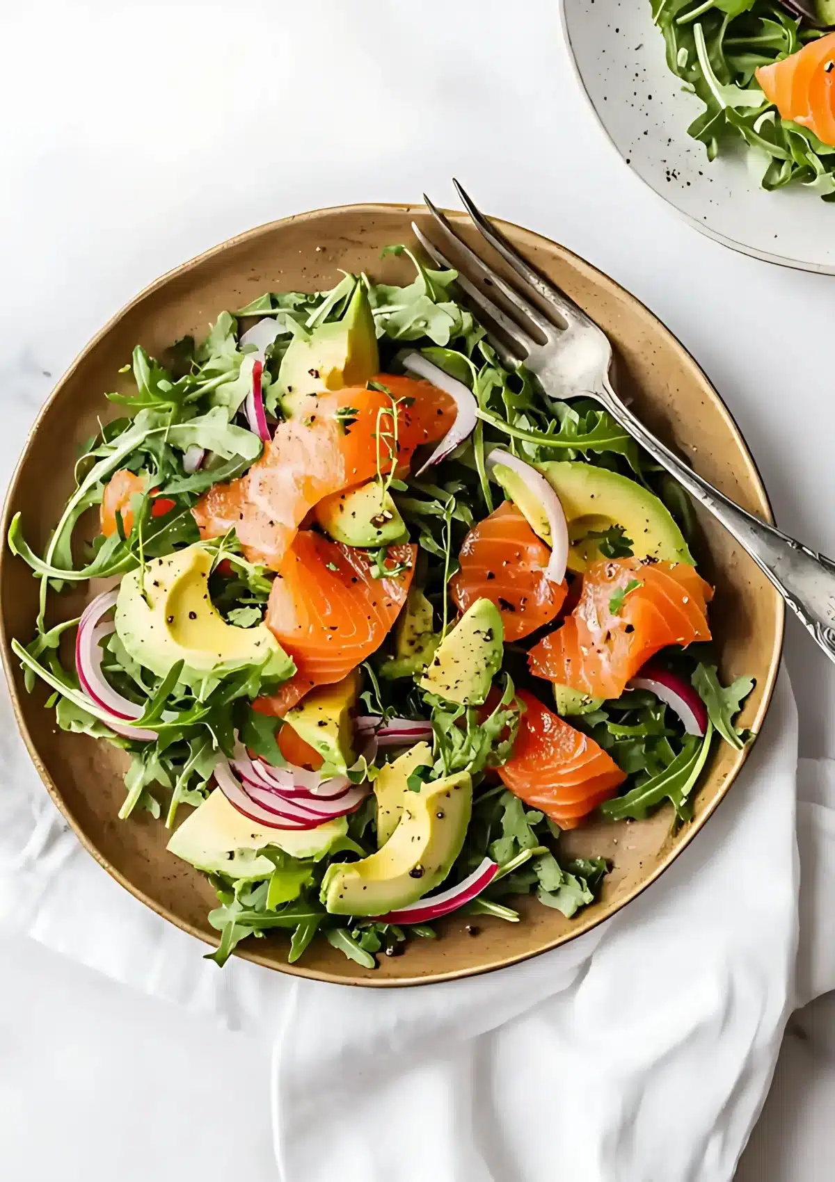 Overhead view of smoked salmon salad in golden ceramic bowl on white marble surface with vintage silverware