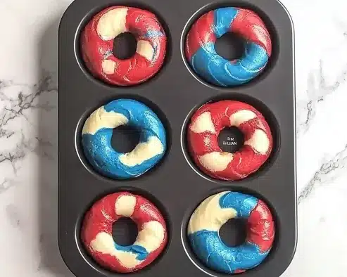 Black donut pan with six baked patriotic cake donuts showing red, white, and blue marbled swirl patterns, overhead view on white marble surface