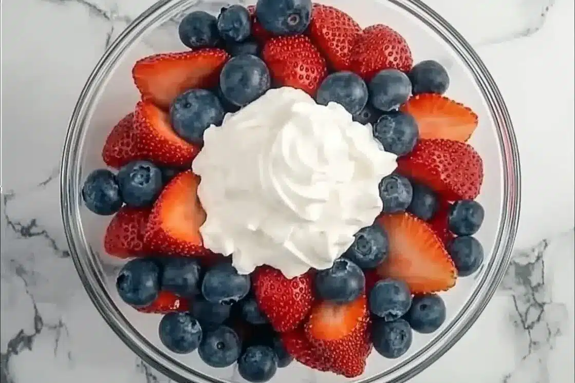 Overhead view of fresh strawberries and blueberries arranged in a circle around white whipped cream center in glass bowl on marble background