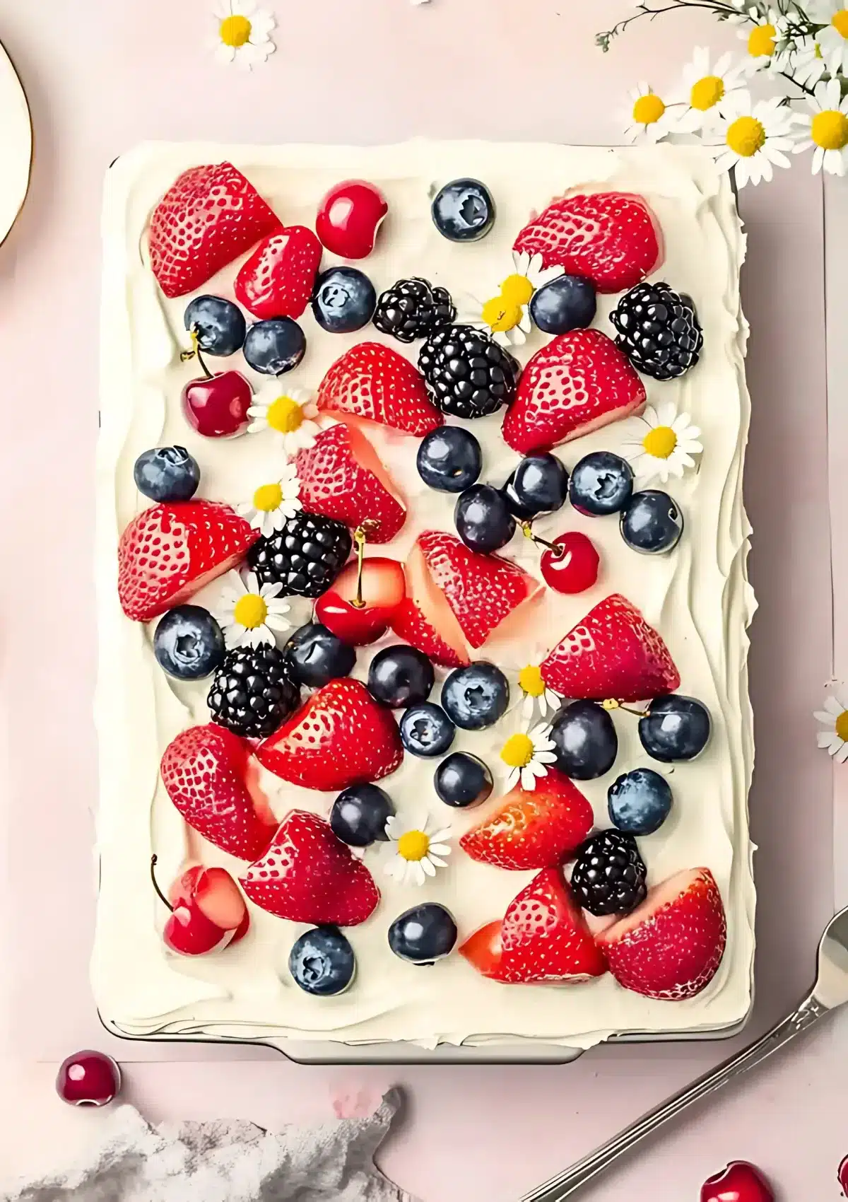 Overhead view of rectangular sheet cake with white whipped cream frosting beautifully decorated with fresh strawberries, blueberries, blackberries, cherries and cheerful daisy flowers on a soft pink background