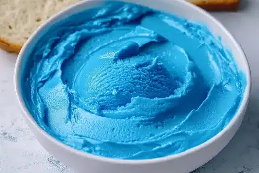 Close-up of a bright blue spirulina-tinted creamy mixture in a white bowl on a light surface with a slice of bread visible at the edge
