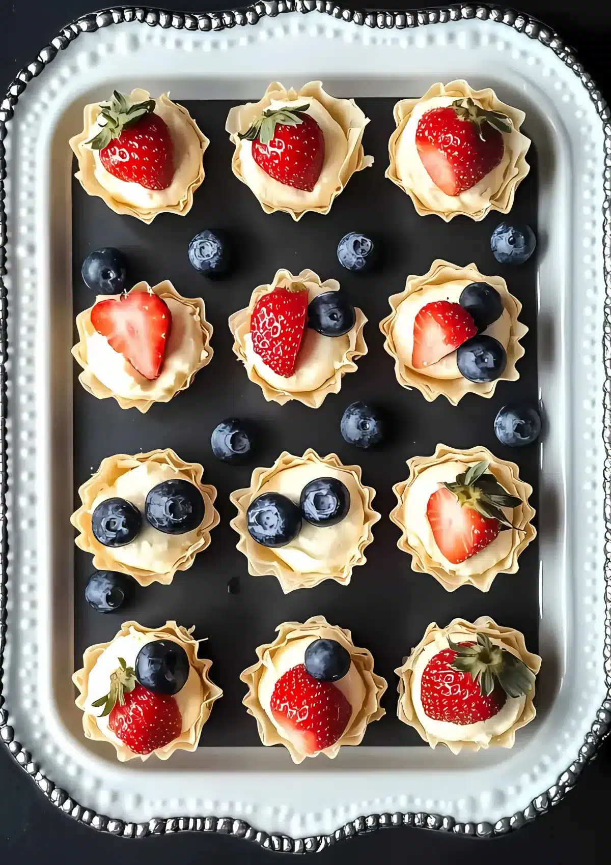 Close-up of cheesecake phyllo cups on silver platter showing detailed berry arrangements with whole strawberries and blueberries on creamy cheesecake filling