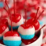 Red, white and blue cherry bombs made with liquor-soaked cherries, white chocolate and blue sugar crystals, arranged in a serving dish.