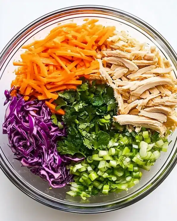 Clear glass bowl with organized sections of Chinese chicken salad ingredients including julienned carrots, shredded purple cabbage, fresh cilantro, chopped green onions, and shredded chicken arranged in neat portions
