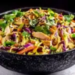 Finished Chinese chicken salad in textured black ceramic bowl topped with sesame seeds and sliced ​​almonds, professional presentation on white surface