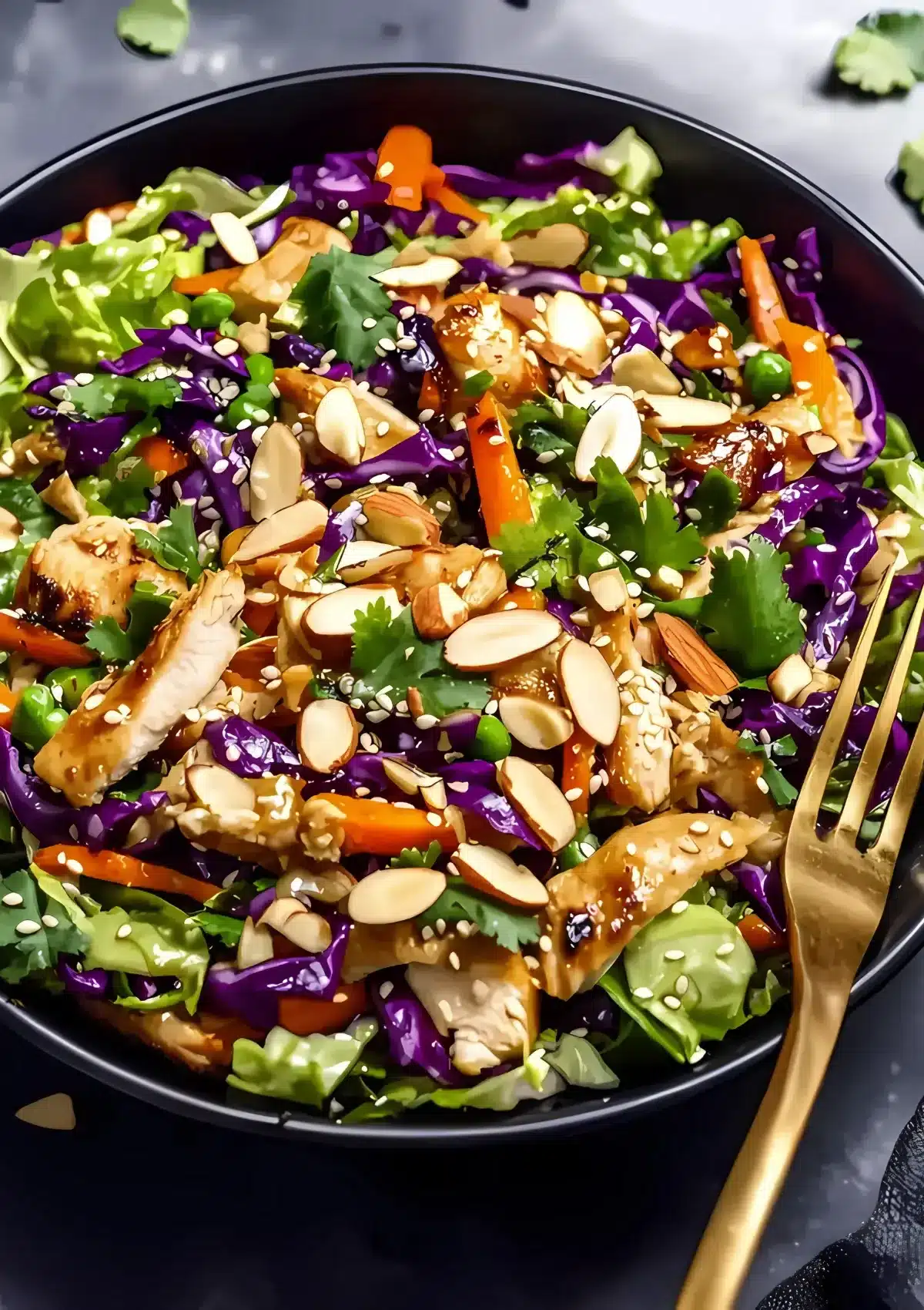Chinese chicken salad in black bowl generously topped with sliced ​​almonds and sesame seeds, gold fork beside bowl, showing rich textures and colors
