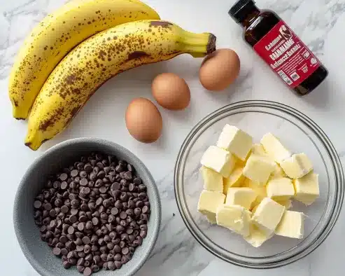 Overhead view of banana bread baking ingredients arranged on white marble surface including overripe bananas, eggs, chocolate chips, butter cubes, and vanilla extract