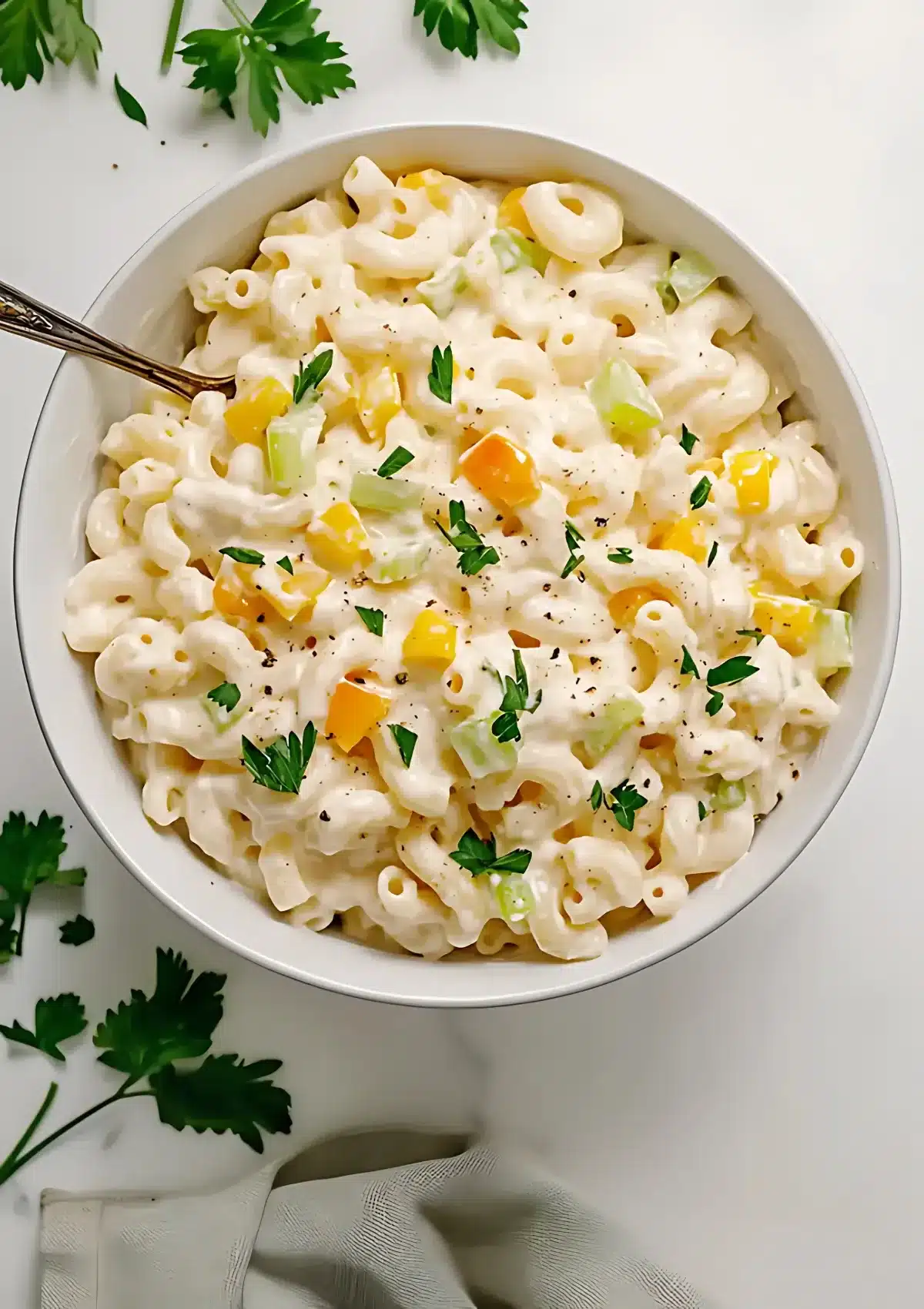 Close-up of creamy macaroni salad in a white bowl showing elbow pasta, colorful bell peppers, celery, and fresh parsley garnish on a marble countertop
