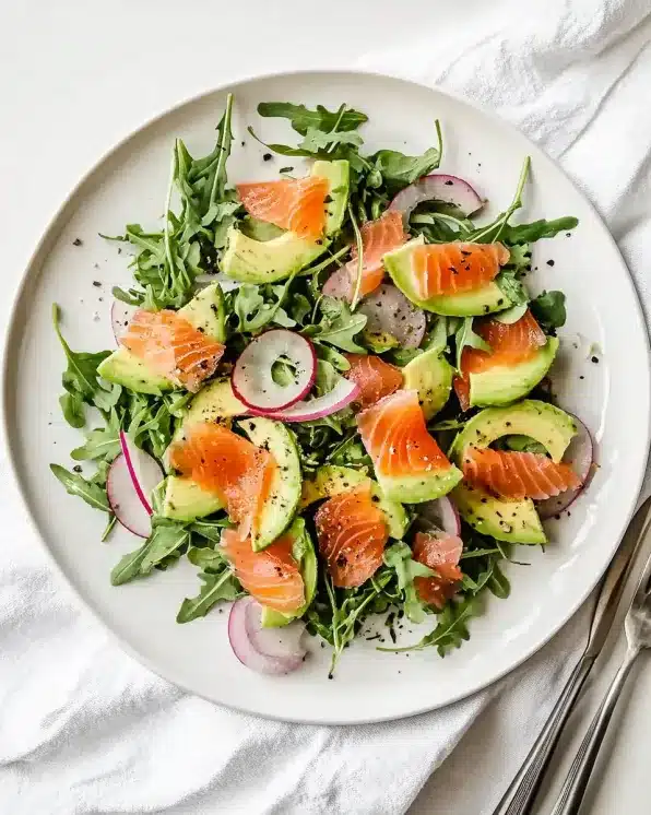 Overhead view of smoked salmon and avocado salad with arugula on white plate with silverware and linen napkin