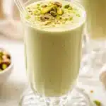 Clear glass of pale green pistachio smoothie with crushed pistachios, with scattered nuts in the background.