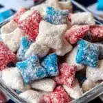 Close-up macro shot of red, white and blue puppy chow pieces in clear glass bowl showing detailed texture of powdered sugar coating on candy-coated rice chex cereal