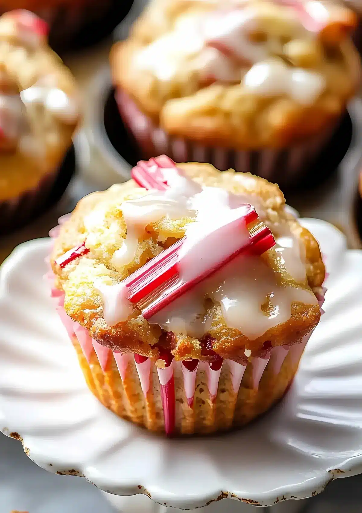 Close-up of rhubarb muffin with sweet glaze drizzled on top and vibrant pink rhubarb chunks, on white scalloped dessert plate