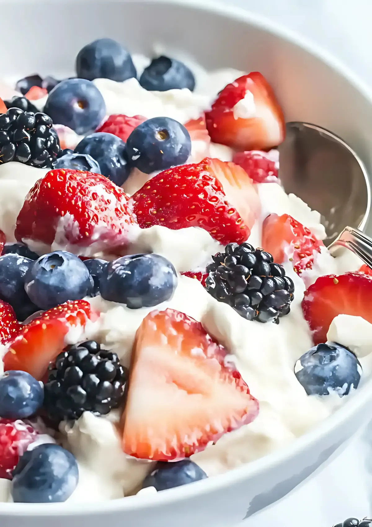White serving bowl filled with triple berry cheesecake fruit salad featuring quartered strawberries, whole blueberries and blackberries in fluffy cream mixture