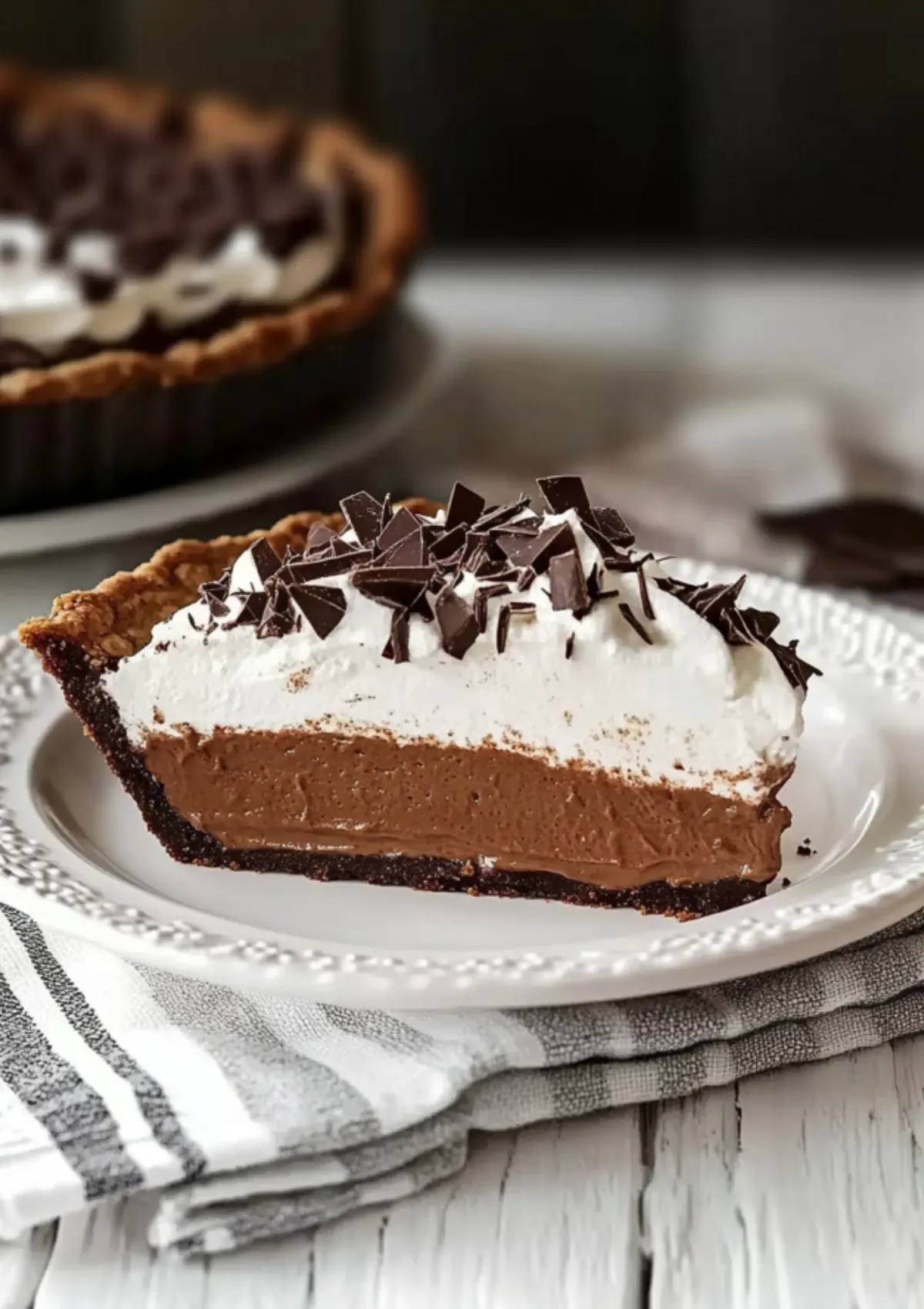 Close-up of chocolate pie slice on decorative white plate with striped napkin and whole pie blurred in background