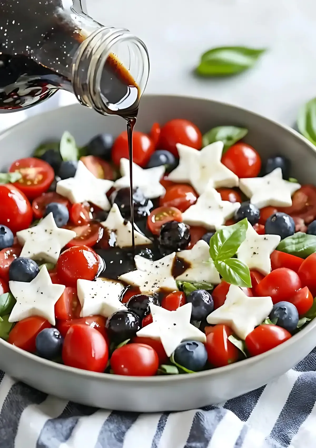 Balsamic vinegar being poured over cherry tomatoes, blueberries, and star-shaped mozzarella cheese with fresh basil leaves