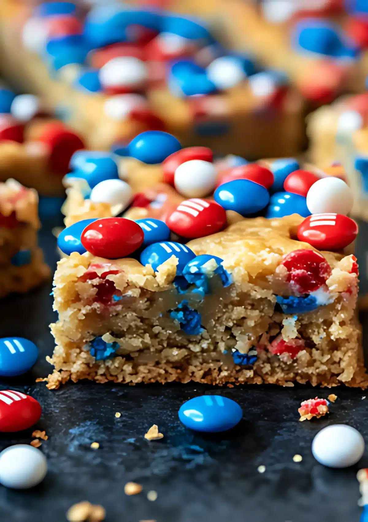 A single Fireworks Blondie bar surrounded by more blondie squares in the background, featuring a caramelized golden top with colorful red, white, and blue M&Ms throughout, on a dark surface