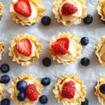 Golden phyllo shells filled with creamy cheesecake and topped with fresh strawberries and blueberries, scattered naturally on parchment for a rustic presentation.