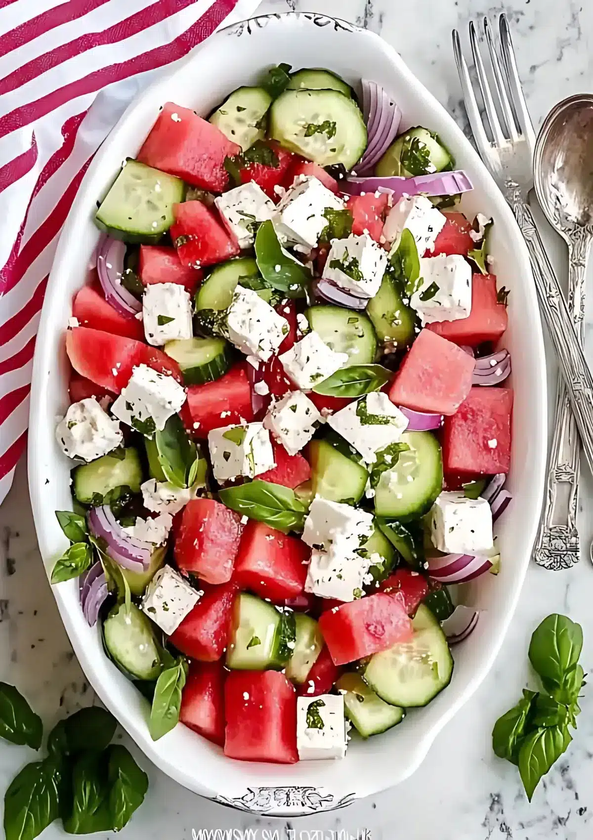 Beautifully plated watermelon feta salad in white oval dish with ornate silverware and pink striped napkin