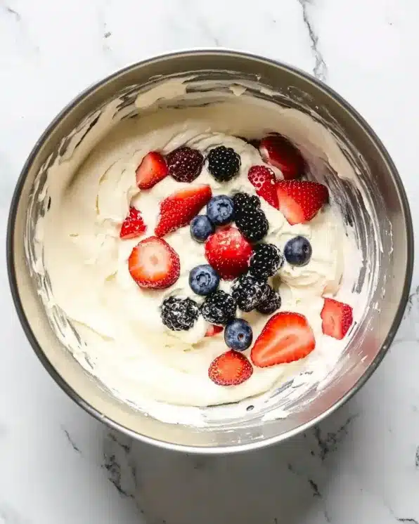 Glass mixing bowl with white cheesecake cream mixture and fresh berries being folded in, showing preparation stage of fruit salad