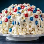 Dome-shaped patriotic popcorn cake on a white scalloped plate featuring white chocolate drizzle over popcorn with red and blue M&Ms against a navy background.