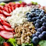 Closeup of Red White and Blue Salad ingredients arranged in sections with sliced strawberries, crumbled feta, plump blueberries and honey glazed walnuts on greens
