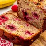 Sliced raspberry banana bread loaf on a wooden board, showing golden crust and moist, fruity interior with vibrant red raspberries.