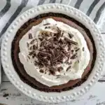 Overhead view of chocolate pie in white ceramic pie dish on striped napkin showing chocolate filling with whipped cream topping and chocolate shavings