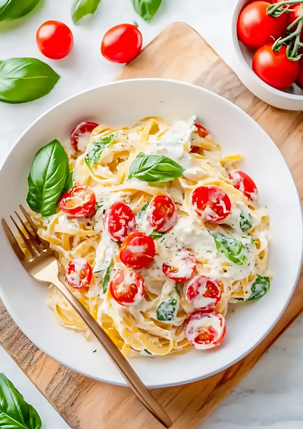 Golden linguine pasta tossed with creamy white ricotta sauce, halved cherry tomatoes, and fresh basil leaves in white ceramic bowl with gold fork, on wooden cutting board