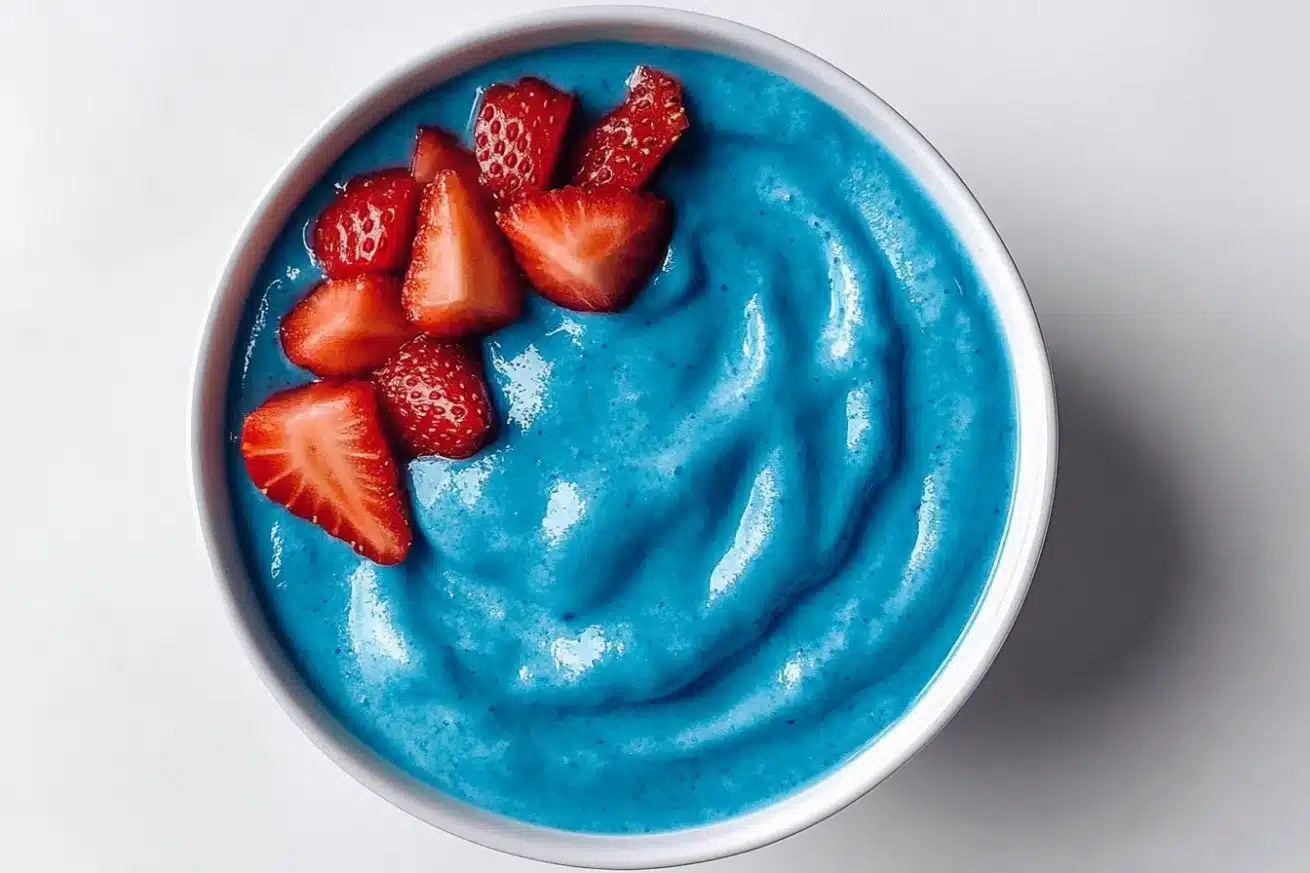 Top-view of a blue spirulina smoothie bowl with fresh strawberry slices arranged on one side in a white bowl on a light background