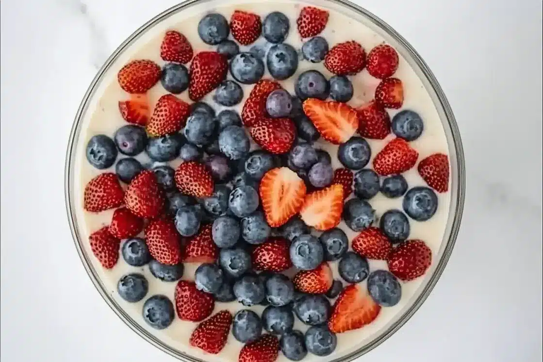 Top-down view of fresh blueberries and sliced strawberries scattered over white chocolate pudding base in glass bowl, creating patriotic red, white and blue colors