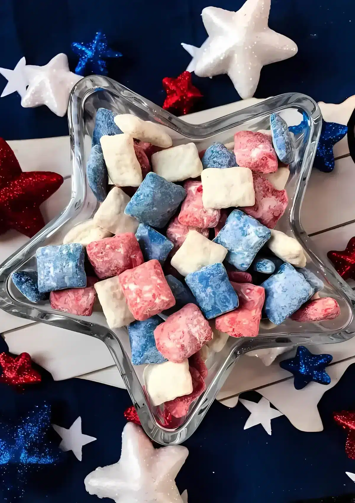 Red, white and blue candy-coated chex cereal mixed together in clear glass star-shaped serving bowl, decorated with foam stars and glittery star decorations on patriotic navy blue background