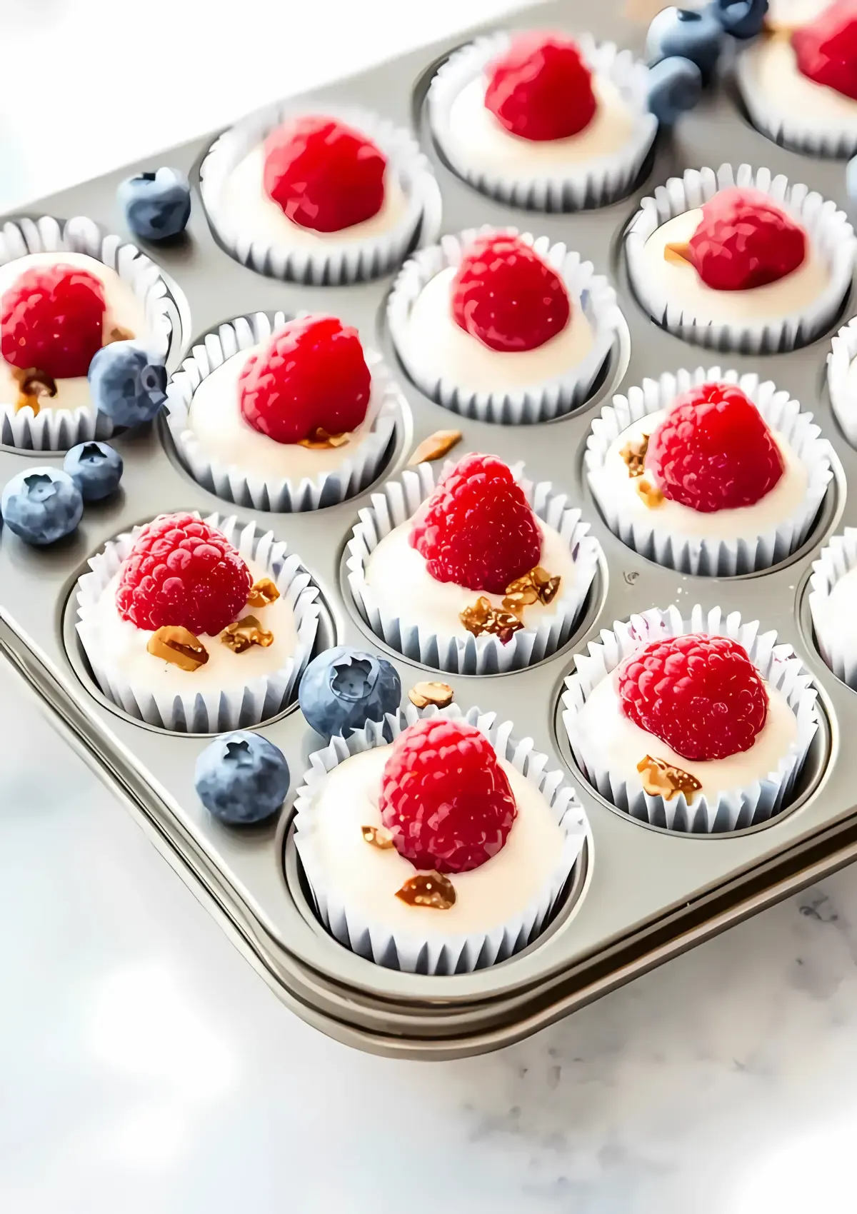 Twelve frozen yogurt bites in white paper liners arranged in a metal muffin tin, each topped with a fresh raspberry and chopped nuts