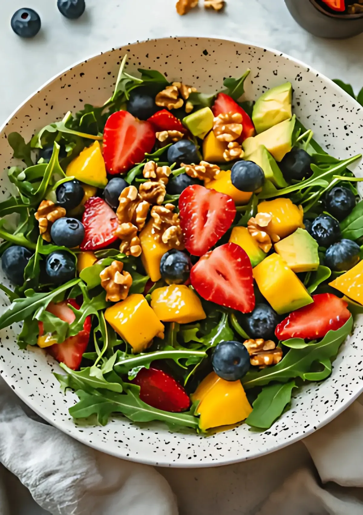 Close-up overhead shot of colorful arugula salad with strawberries, blueberries, mango and walnuts in speckled bowl