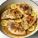 Four golden chicken breasts in enamel braiser with lemon rounds and cooking juices