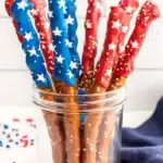 Decorative pretzel rods dipped in red and blue chocolate with white star sprinkles arranged in a mason jar with red and blue sprinkles scattered on the surface and blue napkin in background