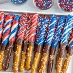 Patriotic chocolate-covered pretzel rods arranged in white wooden tray with red, white, and blue candy coatings and sprinkle decorations, three small glass bowls of sprinkles and blue checkered towel visible